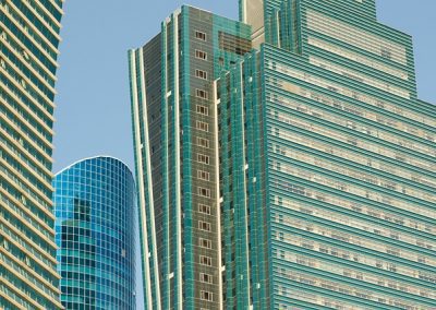 Modern high-rise buildings with glass façades and teal accents rise against a clear blue sky in Kazakhstan. The structures feature various geometric shapes and lines, creating a dynamic urban cityscape.