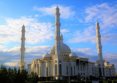 A large white mosque with four tall minarets and blue domes stands under a partly cloudy sky in Kazakhstan, surrounded by greenery.