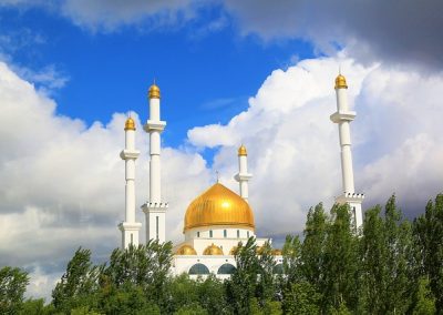 A mosque with a large golden dome and four tall minarets rises above green trees under Kazakhstan’s bright blue sky with dramatic white clouds.