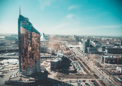 A modern cityscape in Kazakhstan with a tall, glass-fronted skyscraper in the foreground, wide roads, scattered buildings, and a distinctive tent-shaped structure in the background under a clear blue sky.
