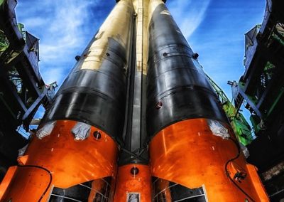A close-up, upward view of a large rocket on a Kazakhstan launch pad, showing its orange and black body, multiple engines at the base, and support structures against a bright blue sky.