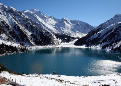 A clear blue lake in Kazakhstan is surrounded by snow-covered mountains and pine trees under a bright, cloudless sky. The snowy peaks are beautifully reflected in the still water.