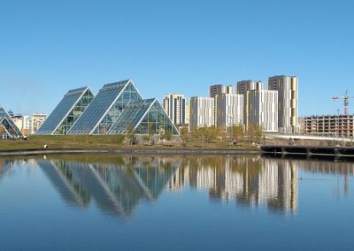 A group of glass pyramid-shaped buildings sits beside a calm lake in Kazakhstan, with modern high-rise buildings and a construction crane in the background under a clear blue sky. The scene is reflected in the water.