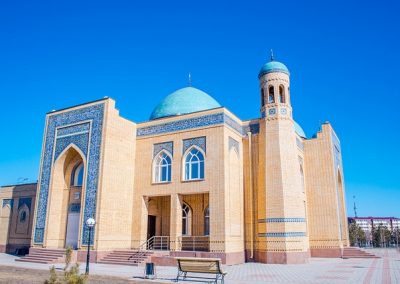 A large mosque in Kazakhstan with beige brick walls, blue-tiled arches, pointed windows, and a tall minaret under a clear blue sky. A turquoise dome crowns the building, with a bench and lamp posts set in front.