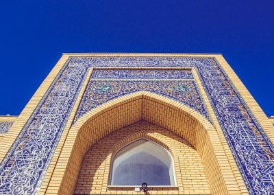 Ornate brick building façade with intricate blue tilework and Arabic calligraphy, featuring a large arched entrance and a pointed window, showcases Kazakhstan's rich architectural heritage under a clear blue sky.