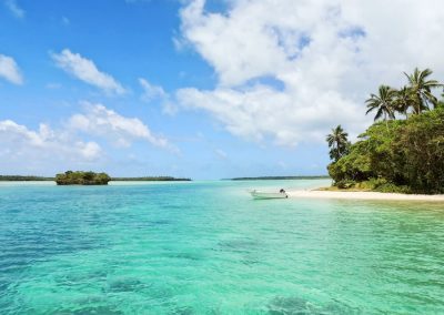 A small white boat rests on the shore of São Tomé and Príncipe, enchanting islands where lush greenery and palm trees meet crystal-clear turquoise water stretching to the horizon beneath a partly cloudy blue sky.