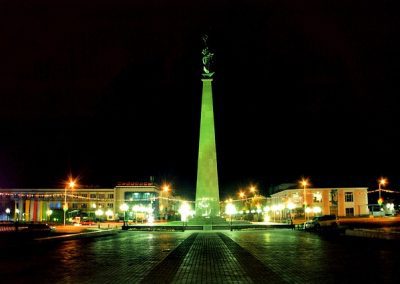 A tall monument with a statue on top stands in a brightly lit square at night in Kazakhstan, surrounded by buildings and glowing streetlights, with a dark sky in the background.