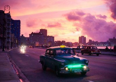 A vintage green taxi cruises along a coastal city road in Cuba at sunset, with a dramatic pink and purple sky, silhouetted buildings, and glowing streetlights in the background.