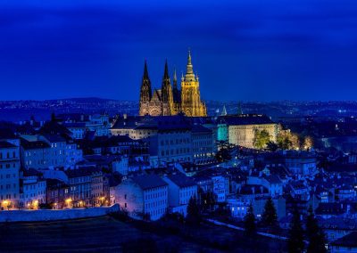 A nighttime cityscape of Prague in the Czech Republic with Prague Castle and St. Vitus Cathedral illuminated, surrounded by dark, blue-lit buildings and streets under a deep blue sky.