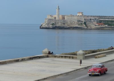 A classic red car cruises along a seaside road in Cuba, with a stone fortress and lighthouse perched on the rocky shore in the background under a clear sky.