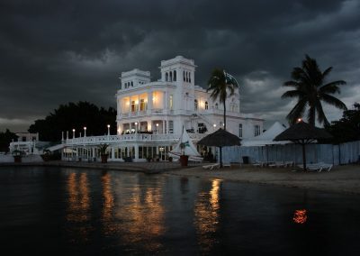 A grand white mansion with glowing lights stands by the water under a dramatic, dark cloudy sky in Cuba. Palm trees, thatched umbrellas, and empty sun loungers line the sandy shore, evoking a serene Caribbean escape.