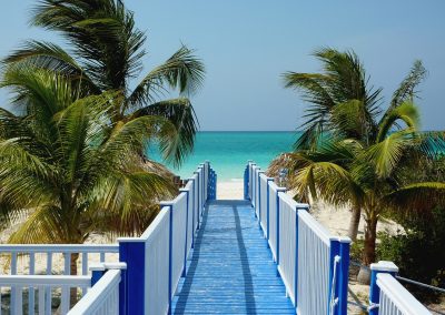A wooden walkway with blue and white railings leads through palm trees to a sandy Cuban beach and turquoise sea under a clear blue sky.