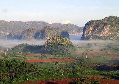 Lush green valley in Cuba with red soil fields, scattered palm trees, and large limestone hills shrouded in morning mist, set against a backdrop of distant mountains under a partly cloudy sky.