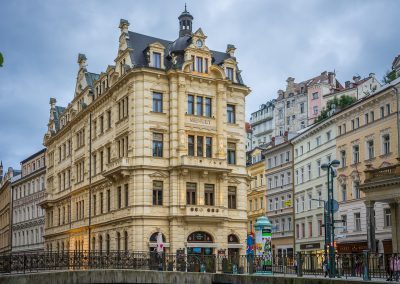 A grand historic building with ornate architectural details stands at a street corner in the Czech Republic, surrounded by other classic European buildings under a cloudy sky. Pedestrians and street lamps line the scene.