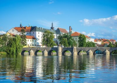 A stone arch bridge crosses a calm river in the Czech Republic, with historic buildings and a church in the background, all under a clear blue sky with some clouds and greenery along the riverbank.