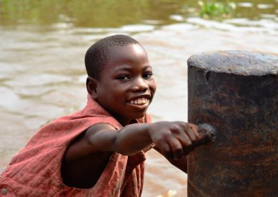 A smiling boy wearing a sleeveless red waistcoat holds onto a large cylindrical metal post near a body of water on a sunny day.