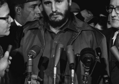 A man with a beard and military-style clothing stands among a group of people, speaking into multiple microphones at what appears to be a press event in Cuba. The setting is in black and white.