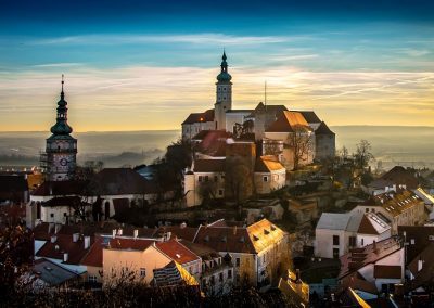 A scenic view of a historic Czech Republic town with red-roofed buildings, two prominent church towers, and a castle on a hill, under a vibrant sky at sunrise or sunset.