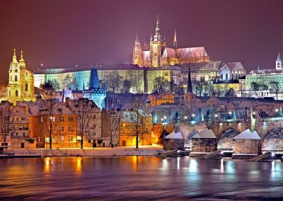 A night-time cityscape of Prague, Czech Republic, featuring illuminated historic buildings, the lit-up Prague Castle complex, snow-covered rooftops, and the Vltava River reflecting the glowing lights.