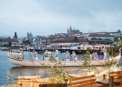 A large white rowing boat with people in matching striped shirts and hats rows on a river in the Czech Republic, with historic buildings and Prague Castle visible in the background under a cloudy sky.