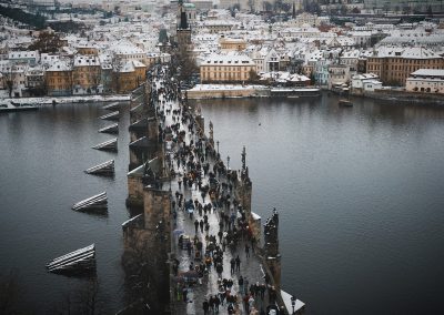 A snowy cityscape in the Czech Republic shows people walking across Prague’s historic Charles Bridge, spanning the Vltava River, with old buildings dusted in snow lining the riverbanks in the background.