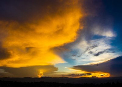 A vibrant sunset sky with dramatic orange, yellow, and blue clouds above a dark landscape and distant hills. The colours blend strikingly, creating a vivid and dynamic scene.