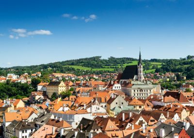 A picturesque Czech Republic town with red-tiled roofs, a prominent church with a tall spire, and green hills in the background under a clear blue sky.