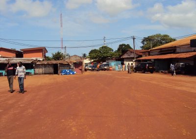 A wide, reddish dirt road runs through a small Gabonese town with scattered buildings, parked cars, and several people walking under a partly cloudy sky. Some trees and utility poles are visible in the background.