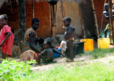 Several children and an adult woman are gathered outdoors near a hut in Guinea-Bissau, with some children standing and others sitting. Plastic containers, a tub, and greenery can be seen in the foreground.