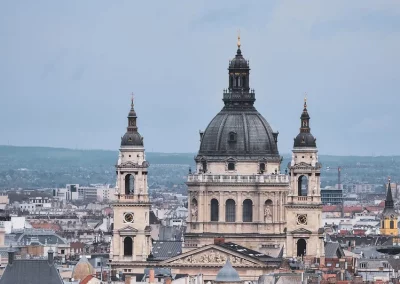 A panoramic view of Budapest, Hungary, featuring St Stephen’s Basilica with its central dome and twin towers, surrounded by historic buildings under a cloudy sky.