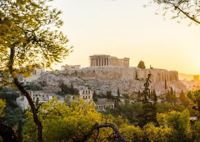 The Parthenon sits atop the Acropolis in Athens, Greece, illuminated by the warm light of sunset, with trees and ancient ruins visible in the foreground.