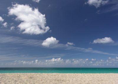 A sandy beach in Jamaica stretches out to a calm turquoise sea under a clear blue sky with scattered white clouds.
