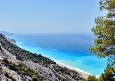 A scenic coastal view in Greece with turquoise blue waters, a sandy beach, rocky cliffs, and lush green trees under a clear blue sky. A large pine tree is visible in the foreground on the right.