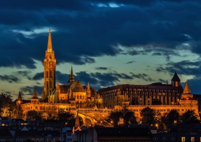 A large, ornate church in Hungary with a tall spire and illuminated golden lights stands against a dramatic evening sky with dark clouds. Trees and adjacent buildings surround the historic structure.