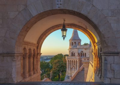 Sunlit stone archway frames a view of Fishermen's Bastion in Budapest, Hungary, with a turret, stairs, and a lantern overhead. Trees and the cityscape are visible in the background under a clear sky.