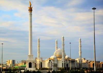 A grand white mosque with multiple domes and tall minarets stands behind a tall white column topped with a golden statue, under a partly cloudy sky in urban Kazakhstan.