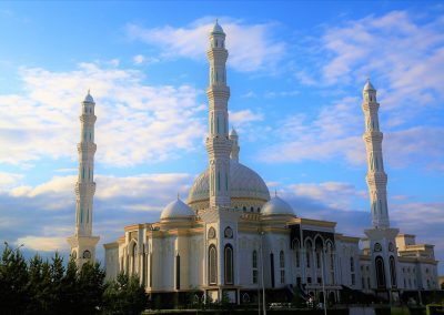 A large, ornate mosque with a central dome and four tall minarets stands against a blue Kazakhstan sky with scattered clouds, surrounded by lush greenery at its base.