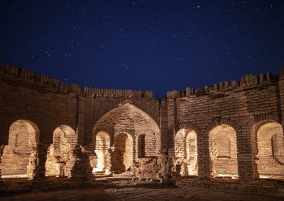 Ancient brick ruins with arched doorways and windows are illuminated at night under a clear, star-filled sky. The structure has weathered walls and an open central area.