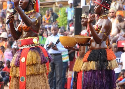Three women in vibrant traditional Guinea-Bissau attire perform a cultural dance outdoors, holding bowls and a knife, as a crowd watches. They wear grass skirts, beaded jewellery, and ornate headpieces.