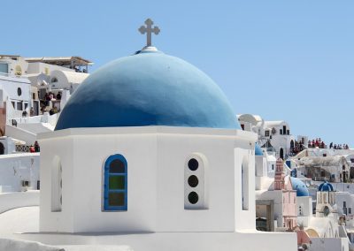 A white building with a blue domed roof and a cross on top, featuring small stained-glass windows, stands among other white structures under a clear blue sky in Santorini, Greece.