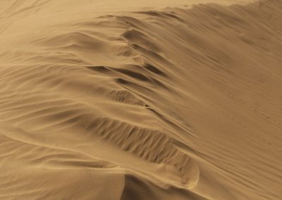 A close-up view of smooth, windswept sand dunes with gentle ridges and shadows, creating a textured, wavy surface in a desert landscape.