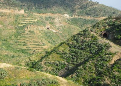 Hilly landscape with green vegetation, terraced slopes, and two visible tunnels carved into the hillsides. A dirt track winds through the terrain under a clear sky.
