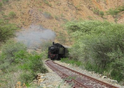A black steam train travels on a winding railway track through green shrubs and rocky hills under a clear blue sky, with smoke rising from its chimney.