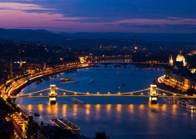 A night-time view of Budapest, Hungary, with the illuminated Chain Bridge spanning the Danube River, city lights reflecting on the water, and historic buildings visible against a twilight sky.