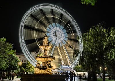 A large Ferris wheel spins at night in Hungary, creating a blurred light effect. In the foreground, a lit statue fountain stands among trees, with people gathered nearby in a vibrant city square.