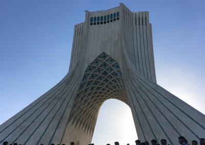 A large group of people stands in front of the Azadi Tower in Tehran, Iran, with a clear blue sky in the background. The monument’s unique geometric arches rise above the crowd.