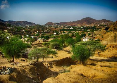 A rural landscape with scattered houses, green trees, and dry, sandy terrain, set against rolling hills under a clear blue sky.