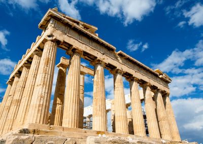 The image shows the ancient Parthenon temple in Athens, Greece, with tall marble columns rising against a bright blue sky and scattered clouds. Some parts of this iconic Greek landmark appear weathered and partially damaged.