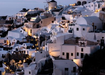 Whitewashed buildings and windmills of Santorini, Greece, glow with warm lights at dusk, set against a soft purple sky and the distant sea—a picture-perfect evening in Greece.