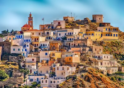 Colourful houses and buildings stack up a sunny hillside in a coastal Greece village, with a church featuring a red dome and clock tower in the background, all beneath a bright blue sky.